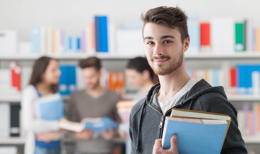 Junger Mann steht mit Büchern in der Hand in einer Bibliothek. Im Hintergrund stehen andere Menschen, ebenfalls mit Büchern in der Hand.