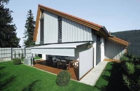 Haus mit Terrasse und Garten. Ueber und seitlich von dieser befinden sich Markisen, die Schatten spenden