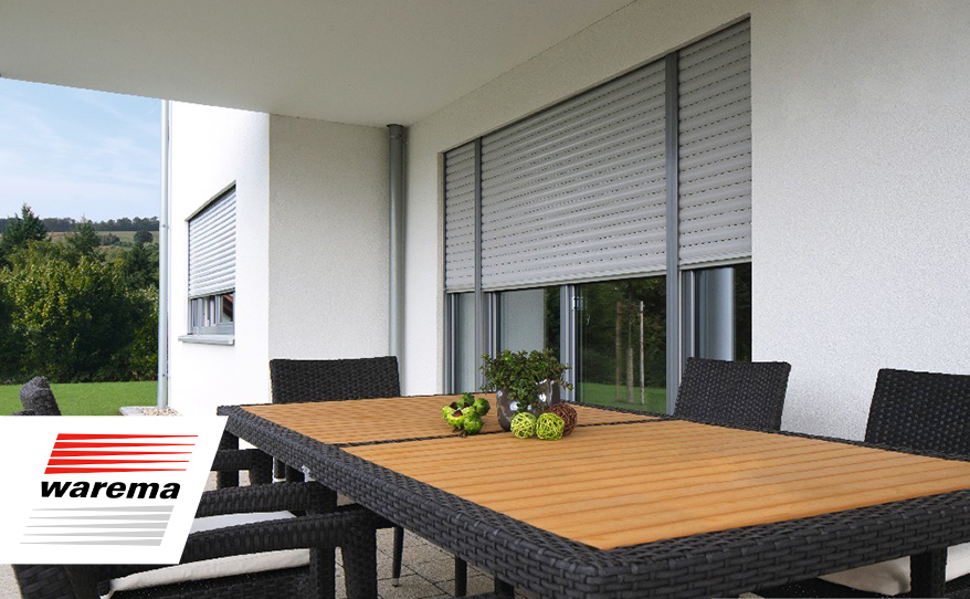 Terrasse mit Tischen und Stühlen. Im Hintergrund Haus mit Fenstern, die zur Hälfte mit heruntergelassenen Aufsetz Rollläden bedeckt sind