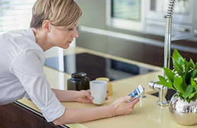 Eine Frau lehnt an einer Küchenzeile und hält ein Smartphone und eine Tasse in der Hand