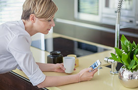 Eine Frau lehnt an einer Küchenzeile und hält ein Smartphone und eine Tasse in der Hand