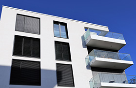 Haus mit drei Etagen und drei Balkonen aus Glas. Die Fenster sind mit Raffstores bedeckt