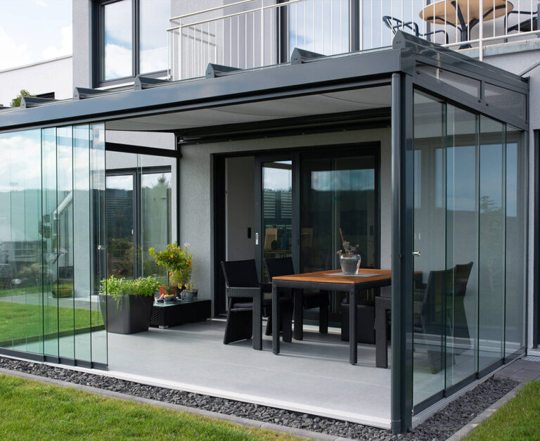 Eine moderne, verglaste Terrasse mit Terrassendächern, ausgestattet mit einem Holztisch, schwarzen Stühlen, Topfpflanzen und Glasschiebetüren, die an ein modernes Haus mit einem darüber liegenden Balkon angebaut ist.