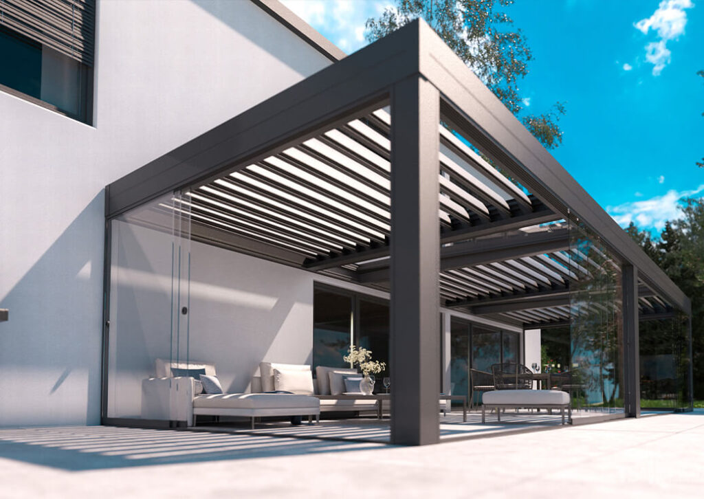 Eine moderne Außenterrasse mit einer eleganten Metallpergola von Terrassendächer, weißen Polstermöbeln, Glaswänden und einer Vase mit Blumen, die an ein modernes weißes Haus angebaut ist. Bäume und der blaue Himmel bilden die perfekte sonnige Kulisse.