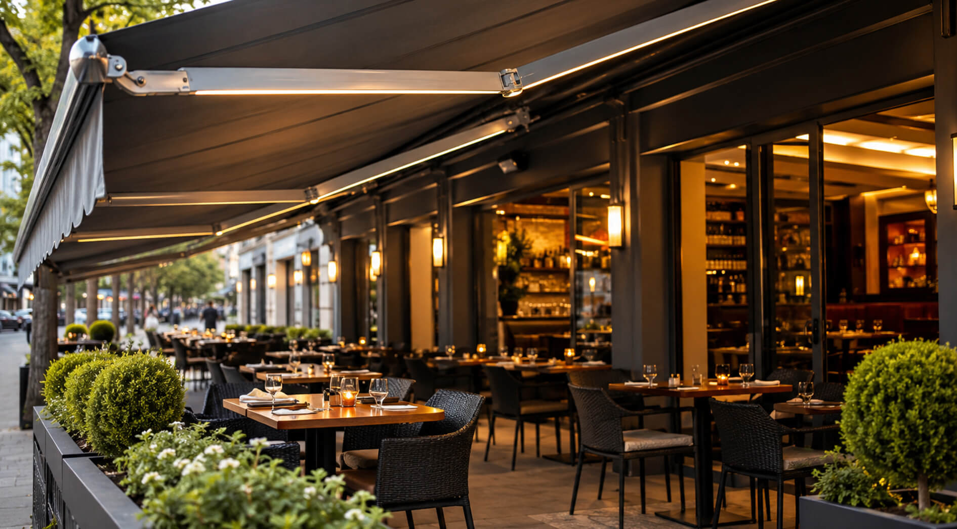 Außenbereich eines modernen Restaurants mit ordentlich gedeckten Tischen, schwarzen Stühlen, Pflanzen und warmer Beleuchtung, geschützt durch eine große Markise von Markisen Gastronomie. Durch die Glastüren ist der Innenbereich des Restaurants zu sehen.