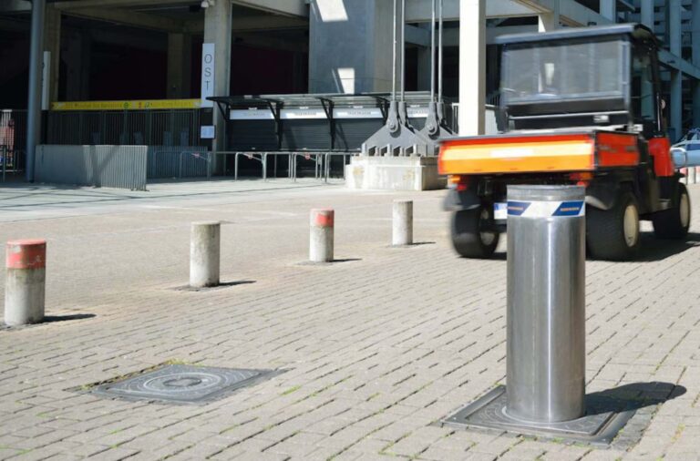 Ein orange-schwarzes Nutzfahrzeug fährt an einer Reihe von Metall- und Betonpoller vorbei, darunter ein versenkbarer Poller, auf einer gepflasterten Fläche in der Nähe eines Gebäudes, hinter dem Metallbarrieren und leere Fahrkartenschalter zu sehen sind.