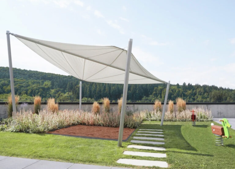 Ein moderner Spielplatz mit einem weißen Sonnensegel über einem Sandkasten, einem Steinweg, Rasen, Pflanzen, einem grünen Federwippgerät und Bäumen, die im Hintergrund unter einem klaren Himmel zu sehen sind.