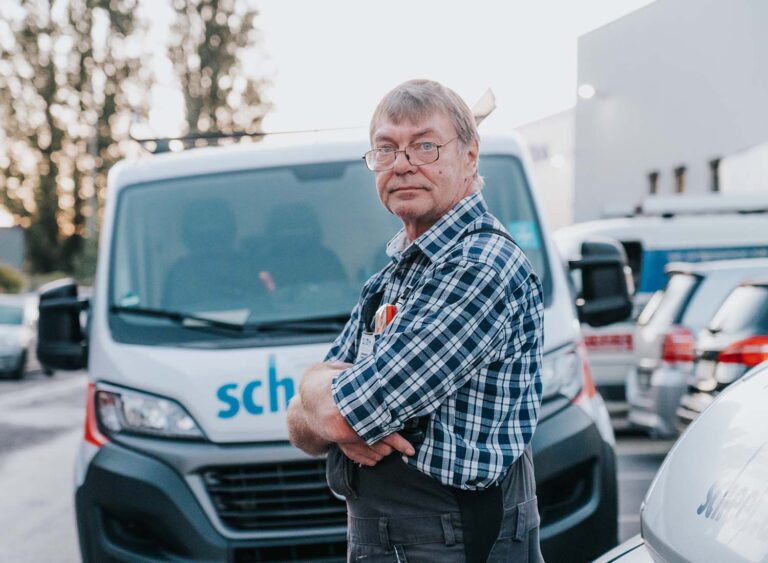 Ein älterer Mann mit Brille und Karohemd steht mit verschränkten Armen vor einem weißen Transporter auf einem Parkplatz und blickt in die Kamera – eine selbstbewusste Ausstrahlung, die auf jahrelange Berufserfahrung hinter dem Steuer hindeutet.