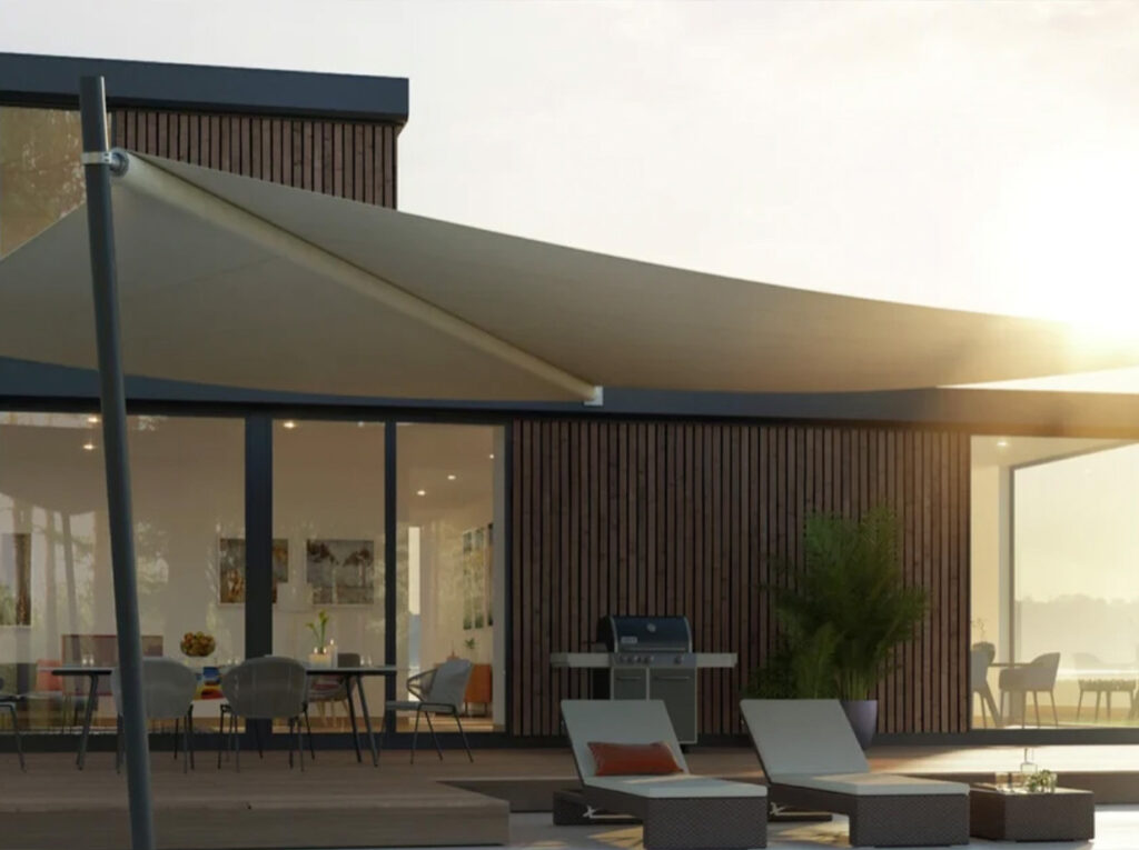 Moderne Terrasse mit Liegestühlen, einem Grill und Tischen unter einem großen, beigen Sonnensegel. Das Gebäude besticht durch Holzvertäfelungen, große Glasfenster und hochwertige Produkte, die für eine warme, einladende Atmosphäre sorgen.