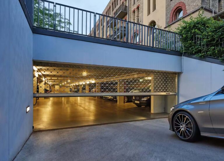Durch massive Rolltore und Rollgitter ist eine moderne Tiefgarage mit mehreren geparkten Autos zu sehen. Vor der Garage steht ein silbernes Auto, und über dem Garageneingang sind Wohnhäuser zu erkennen.