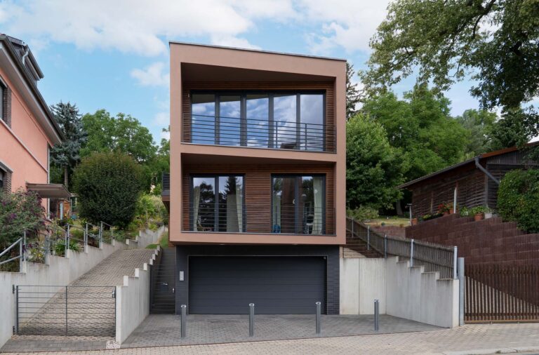Ein zweistöckiges, modernes Haus mit brauner Fassade, einer Garage für zwei Autos auf Straßenebene, die durch versenkbare Poller gesichert ist, sowie Balkonen auf jeder der oberen Etagen.