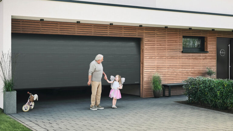 Ein älterer Mann und ein junges Mädchen mit Feenflügeln halten sich vor einer modernen Garage mit Terrassendach in Bochum an den Händen.